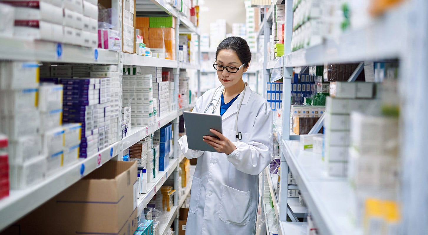  Female pharmacist in a white coat and glasses using a tablet while standing in the aisle of a well-stocked pharmacy warehouse or storeroom.